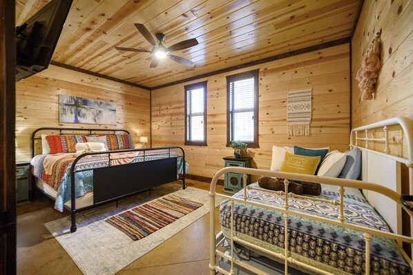 A cabin-style bedroom with two beds, wooden walls, a ceiling fan, and a large window.