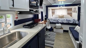 A compact RV kitchen with a sink, stove, and microwave. There is a bed with a blue and white patterned bedspread and a small seating area with a bench and cushions.