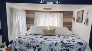 A bedroom with a bed featuring a nautical-themed bedspread, a 'welcome' pillow, and a headboard with anchors. There is a small bedside table with a lamp, a blue bucket, and a basket.