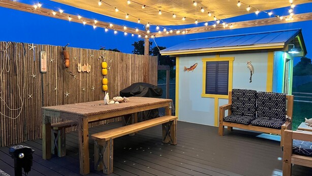 An outdoor deck with a wooden table and benches, string lights, and a small building with a fish mural.