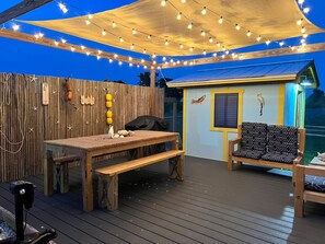 An outdoor deck with a wooden table and benches, string lights, and a small building with a fish mural. - Cozy RV in Homestead, near the Everglades National Park & the Florida Keys! (Homestead)