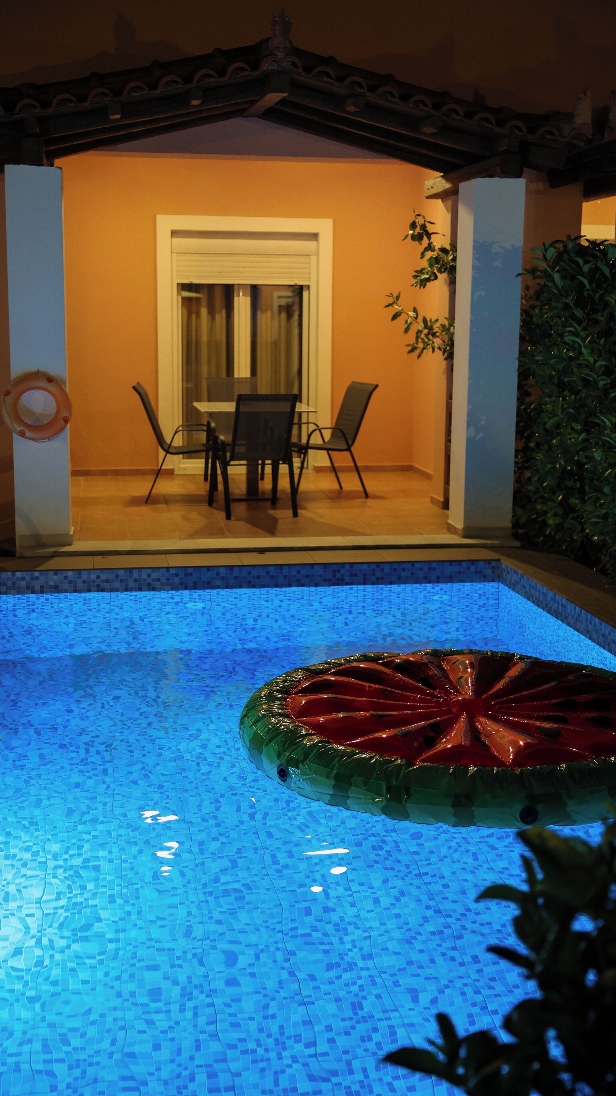A swimming pool with a green inflatable ring, a dining area with chairs and a table, and a door leading to a covered patio.