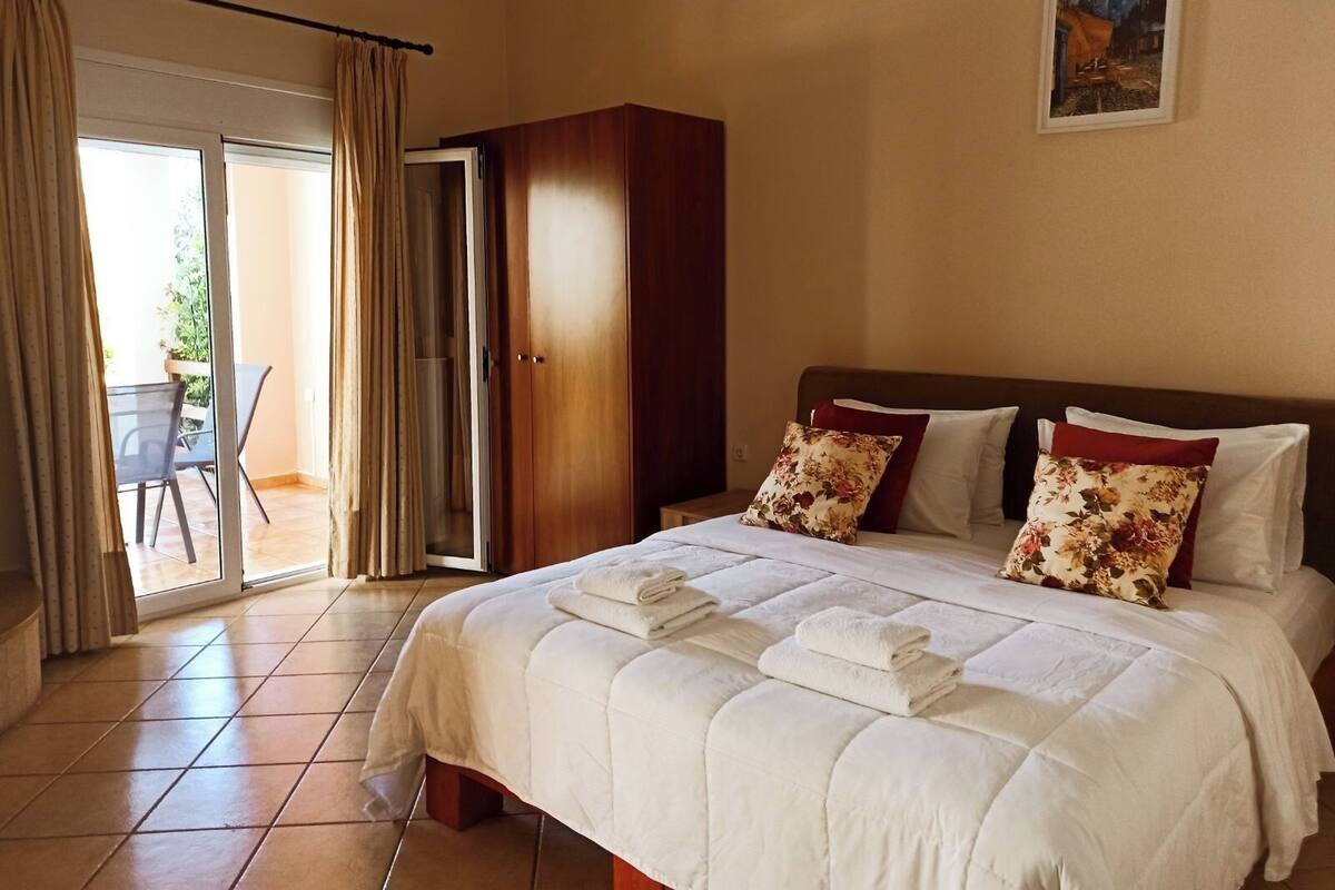 A hotel room with a bed, a wooden wardrobe, a balcony with a table and chairs, and a painting on the wall.