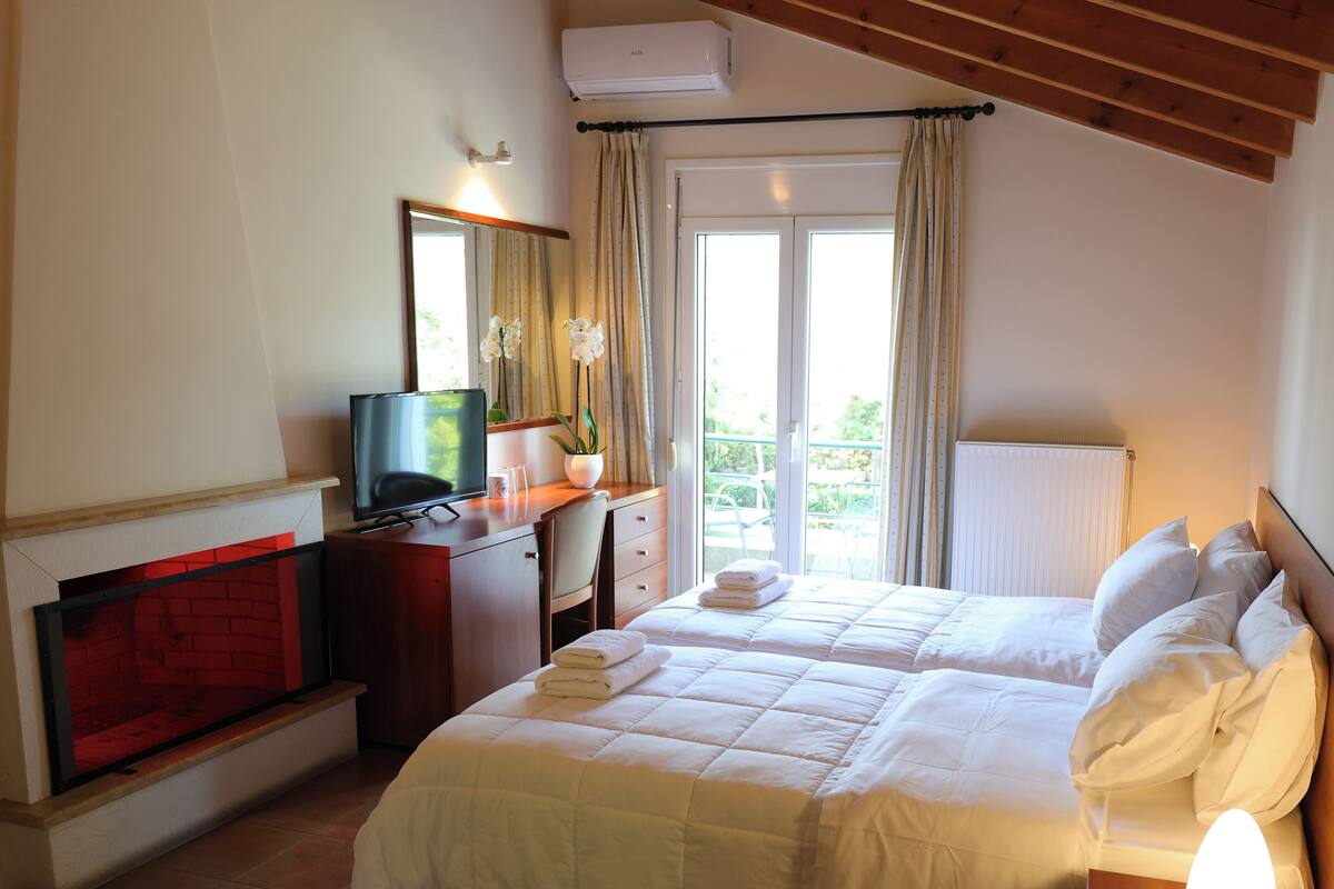 A hotel room with a bed, a TV, a desk with a chair, a fireplace, and a window with curtains.