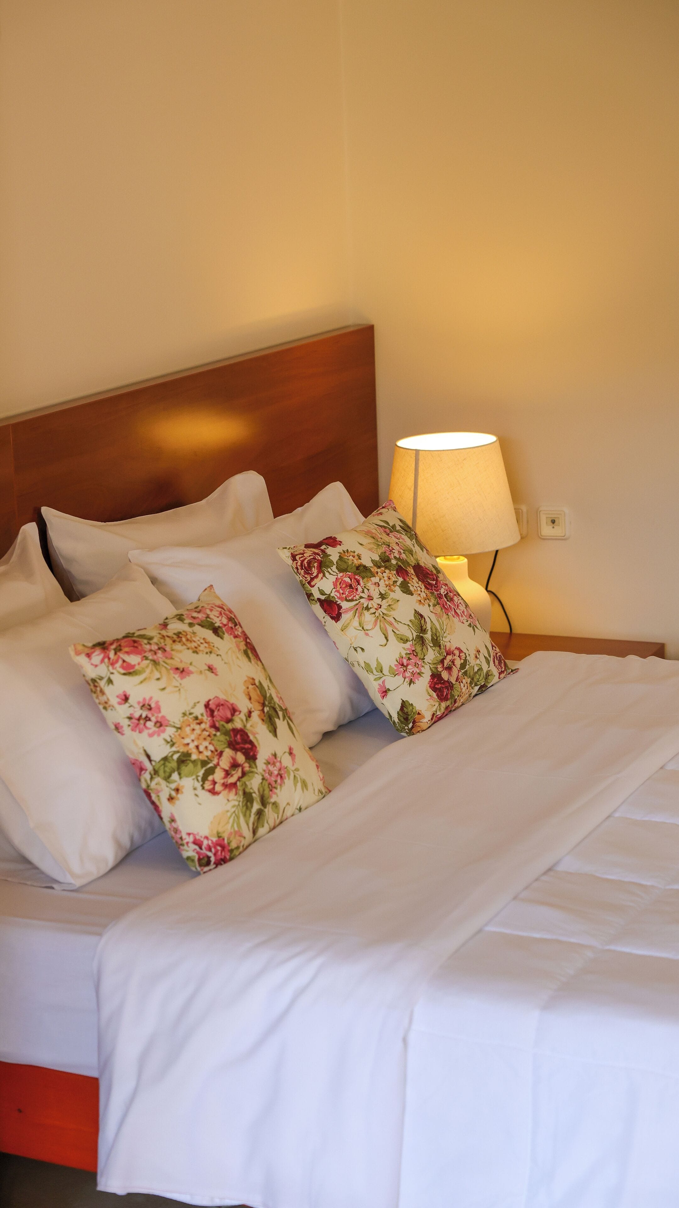 A neatly made bed with white bedding and floral-patterned pillows, a wooden headboard, and a bedside lamp.