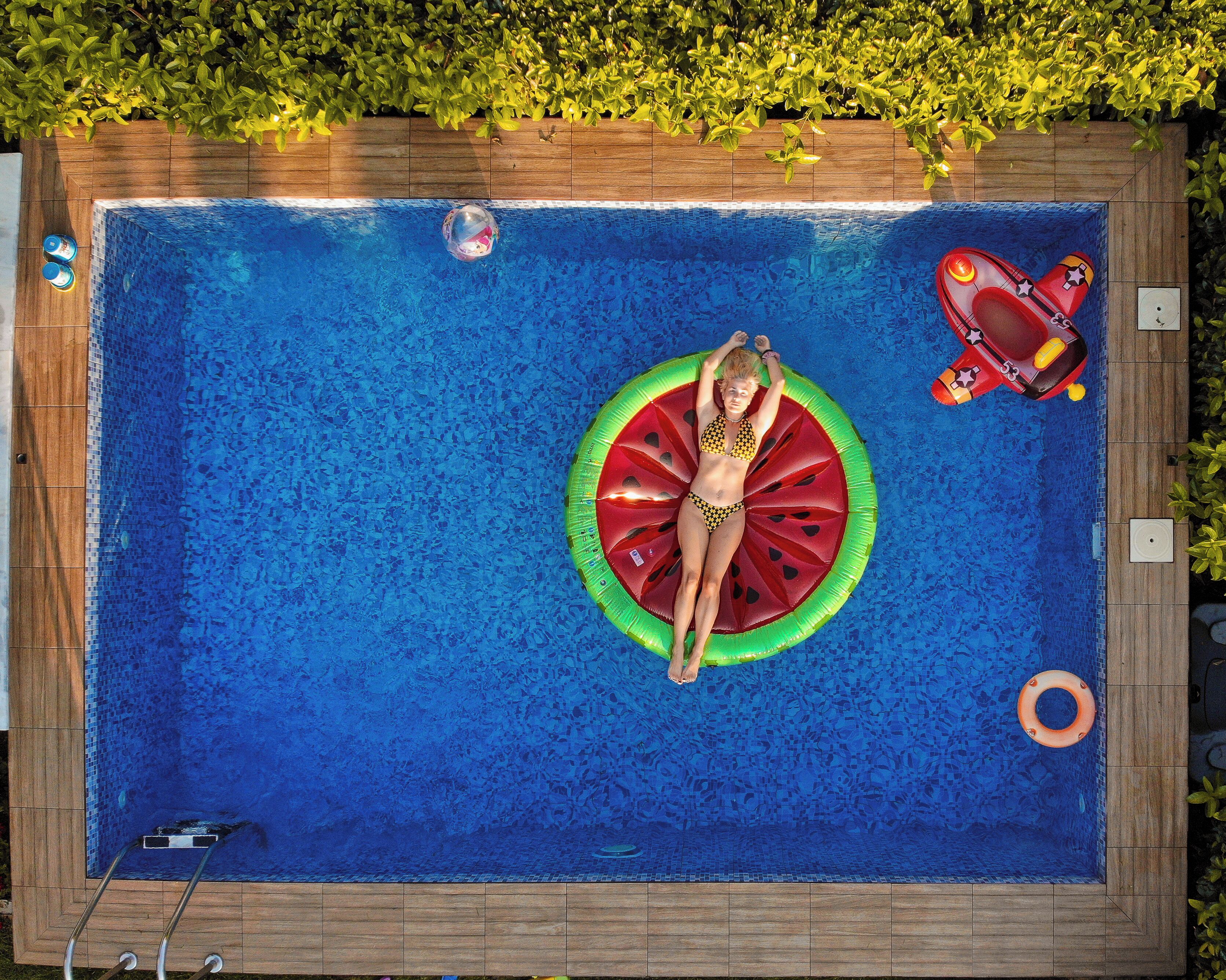 A swimming pool with a person lying on a watermelon float, surrounded by a wooden deck and greenery.