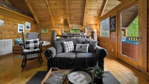 Un interno accogliente con soffitto in legno, un divano nero con cuscini decorati, una poltrona a dondolo e un tavolo da pranzo.