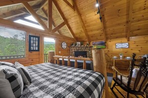 Une pièce chaleureuse aménagée dans un chalet en rondins, avec un lit, une cheminée en pierre, une table à manger et une vue sur l’extérieur.
