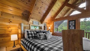 Camera da letto in una baita di legno, con copriletto a scacchi, comodino con lampada e finestra con persiane.