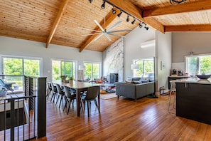 Een ruime woonkamer met houten plafond, een eettafel met stoelen, een bank en een keuken met groot raam.
