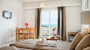 A hotel room with a bed, a desk, a chair, a table with a vase and candles, and a view of the sea. - Sunny Hill Hotel Apartments (Chlorakas)