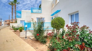 A residential building with white walls and blue accents, a paved walkway, and various plants. - Sunny Hill Hotel Apartments (Chlorakas)