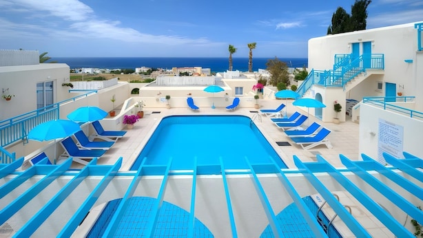 A swimming pool with blue chairs and umbrellas, a view of the sea, and a white building with blue accents. - Sunny Hill Hotel Apartments (Chlorakas)