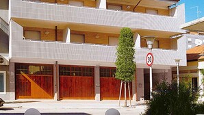Un edificio de varios pisos con un diseño arquitectónico moderno, que presenta una entrada destacada con una puerta roja, una señal de límite de velocidad visible y un jardín bien cuidado con macetas de piedra esféricas.