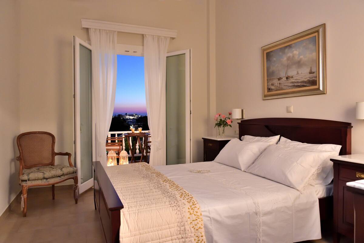 Une chambre d’hôtel avec un lit, une chaise, une table de chevet, un tableau et une vue sur la ville au crépuscule.