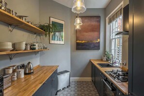 Une cuisine avec des plans de travail en bois, une cuisinière à gaz, un évier et un petit réfrigérateur. On y trouve des étagères remplies de vaisselle et une plante en pot.