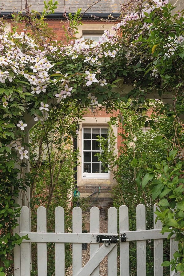 Une porte en bois peinte en blanc, avec une fenêtre derrière, entourée d’une végétation luxuriante et de plantes en fleurs.