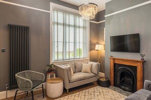 Un salon comprenant un canapé, un fauteuil, une cheminée, un téléviseur et une fenêtre avec des stores.