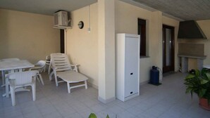 Un petit patio clos avec une table et des chaises blanches, un réfrigérateur, une cuisinière à gaz et une plante en pot.