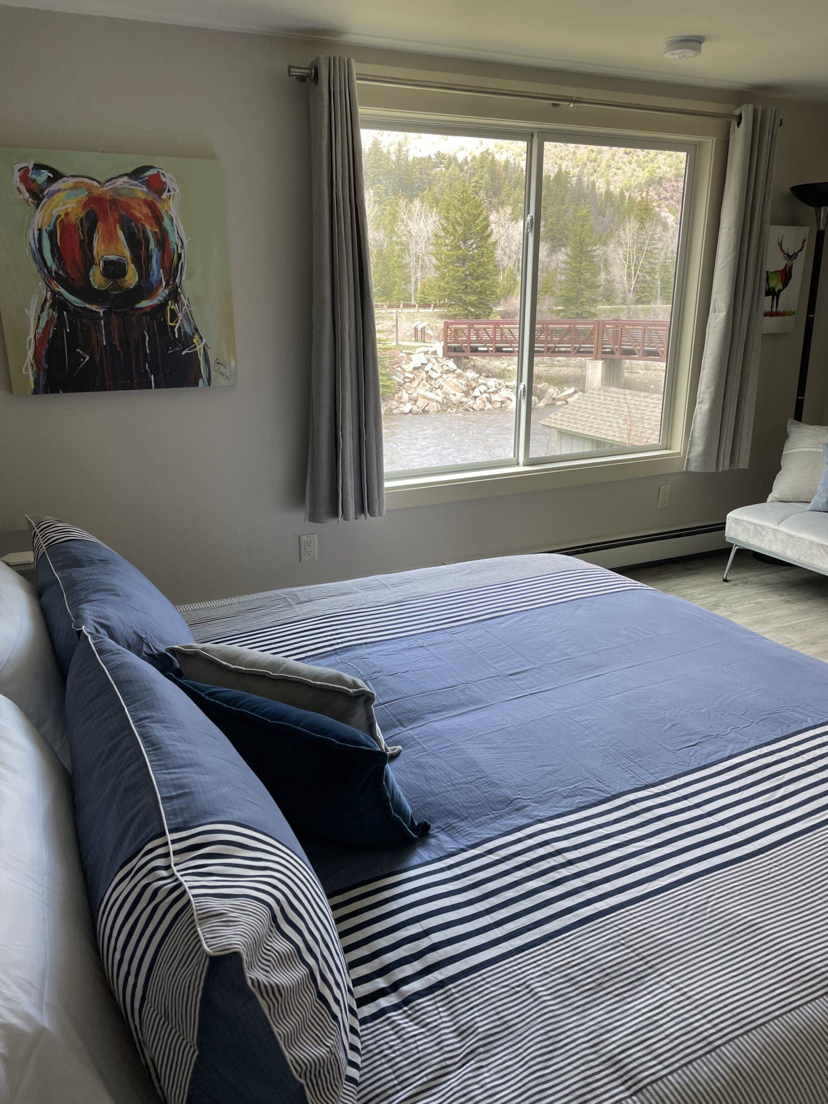 A bedroom with a large bed, a painting of a bear, a window with a view of trees and a bridge, and a chair.