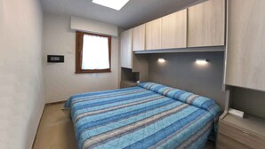 A bedroom with a bed, a window, built-in cabinets, and a ceiling light. - Holiday apartment mit Klimaanlage und Balkon (Lignano Sabbiadoro)