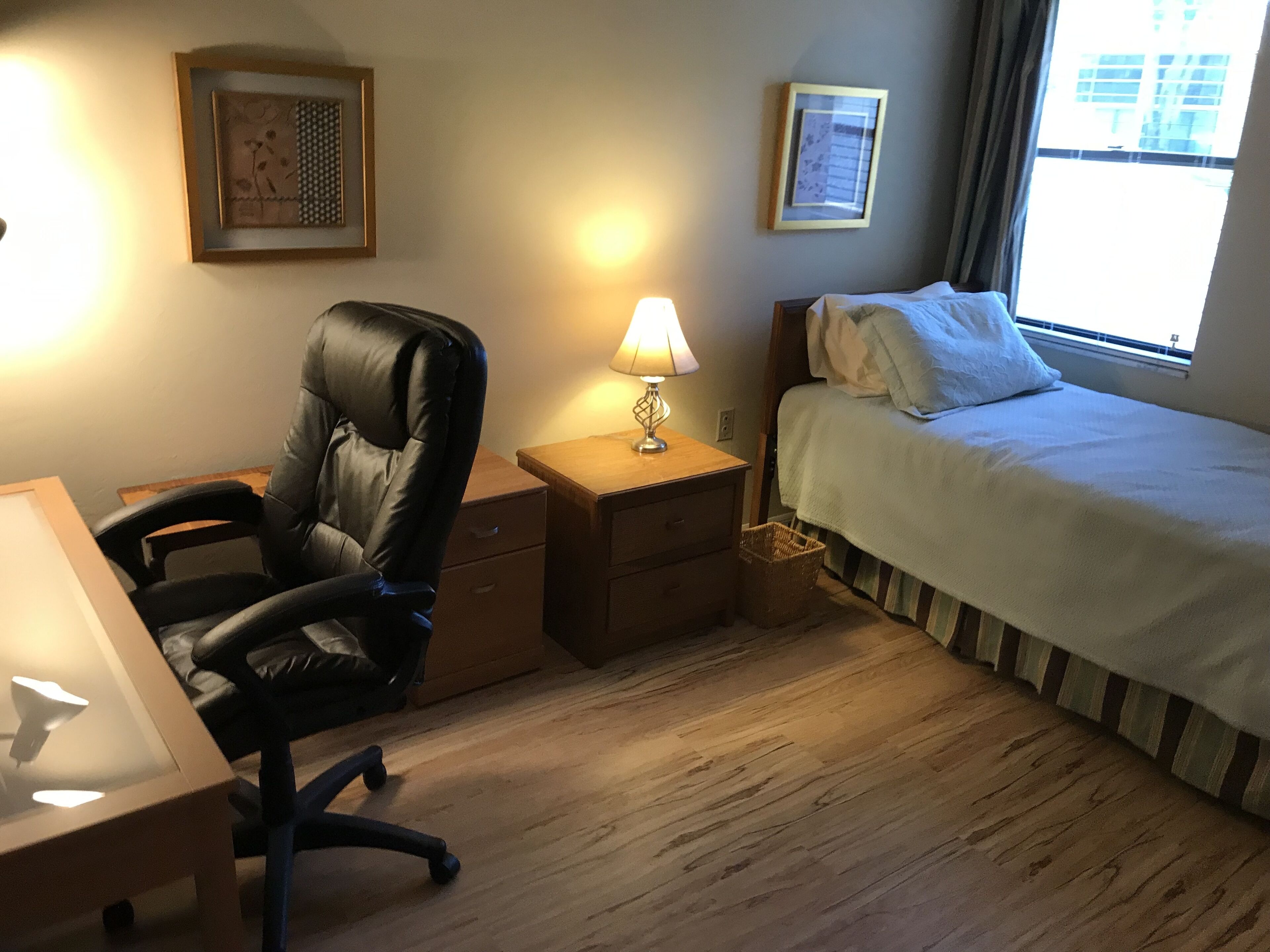 A hotel room with a bed, a desk, a chair, a nightstand with a lamp, and two framed pictures on the wall.