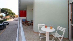 Un balcón con una mesa blanca y dos sillas blancas, una puerta roja, y vistas a coches aparcados y árboles.