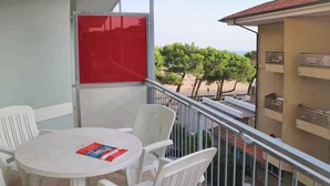 Un balcón con una mesa redonda y sillas blancas, con vista a una playa con árboles y cielo despejado.