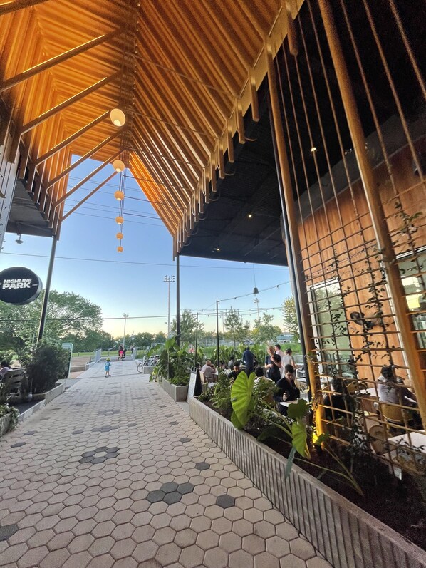 Enjoy dining under a unique architectural design with a geometric ceiling and hanging lights, surrounded by lush greenery and a paved pathway.