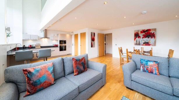Ein modernes Wohnzimmer mit grauen Sofas und farbenfrohen Kissen, einem Holzessttisch und einer Küche im Hintergrund.