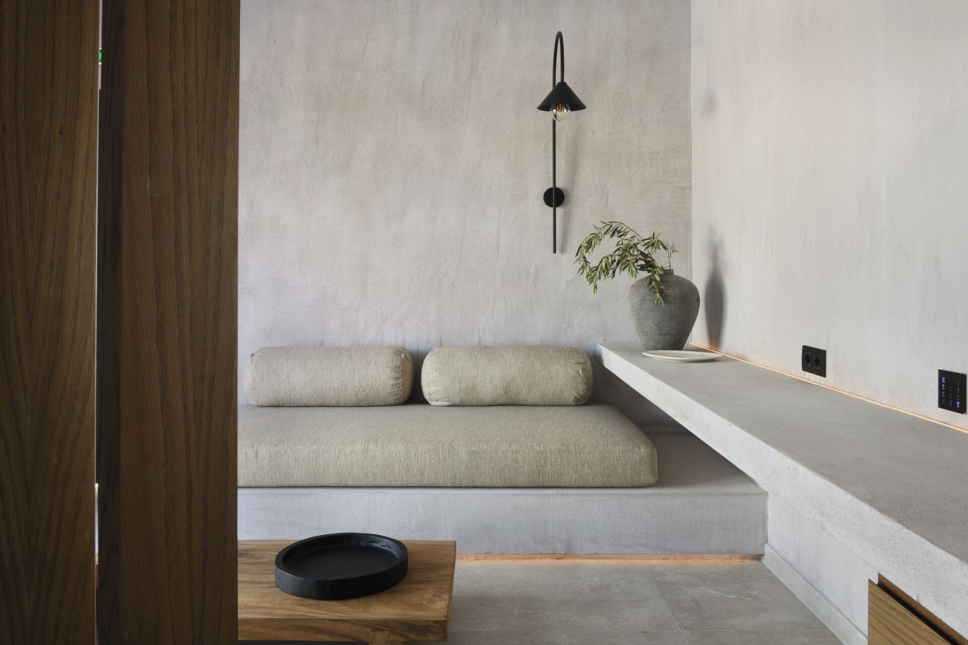 Una habitación minimalista con un banco empotrado, una mesita con una bandeja negra, una lámpara de pared y una planta en maceta.
