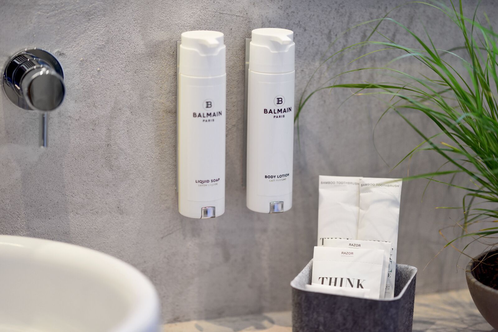 Une salle de bain avec un lavabo blanc, deux distributeurs de lotion pour le corps Balmain blancs, une brosse à dents en bambou et une poubelle portant l'inscription « THINK ».