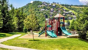 Un parc infantil amb tobogans i elements per escalar, envoltat d'una zona verda i arbres, amb cases al fons.