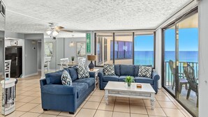 A spacious living room with a large window overlooking the ocean, a ceiling fan, and a dining area with a table and chairs.