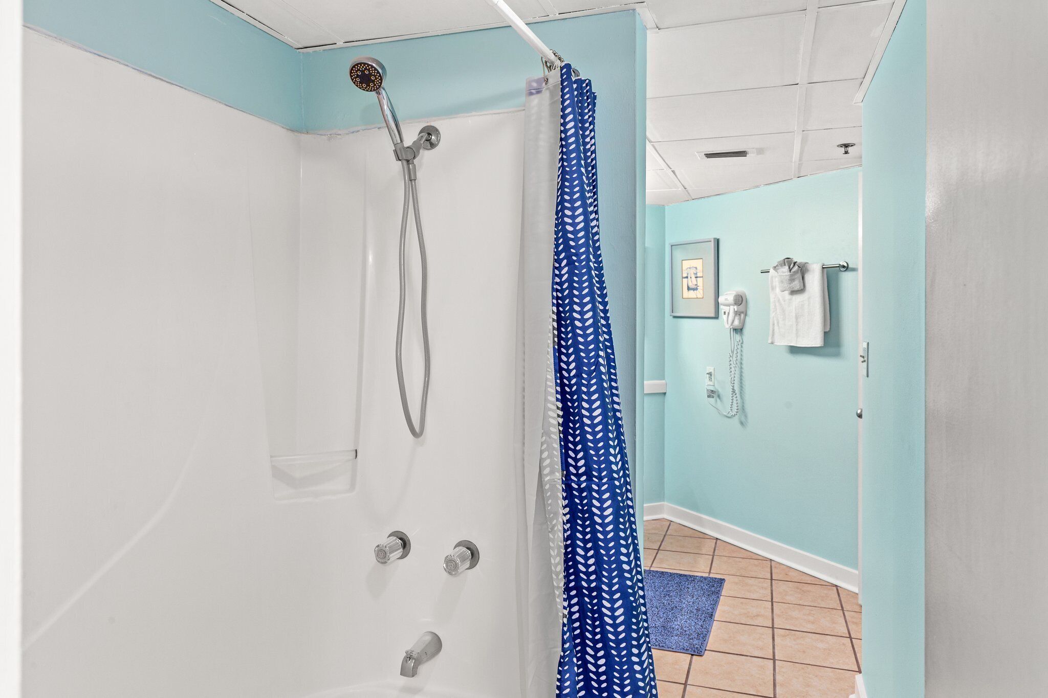 A bathroom with a shower, a blue patterned shower curtain, a white towel rack, and a framed picture on the wall.