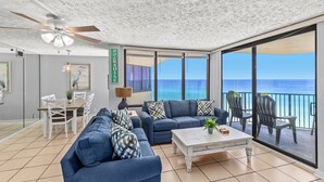 A spacious living area with a balcony overlooking the ocean, featuring a ceiling fan, a dining table with chairs, and a sofa set.