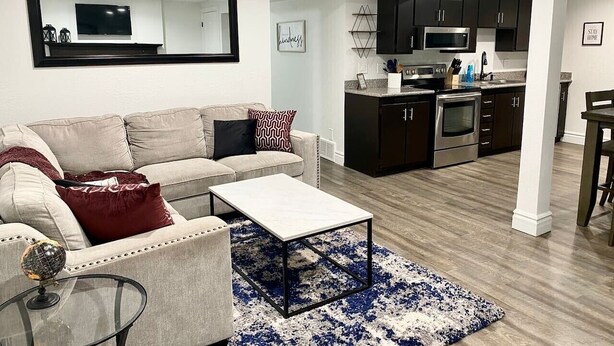 A living room with a sofa, coffee table, and a rug. There is a kitchen area with cabinets and appliances.