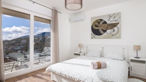 Une chambre avec une grande fenêtre, un lit recouvert d’une couette à motifs floraux, deux tables de chevet avec des lampes et un tableau abstrait accroché au mur.