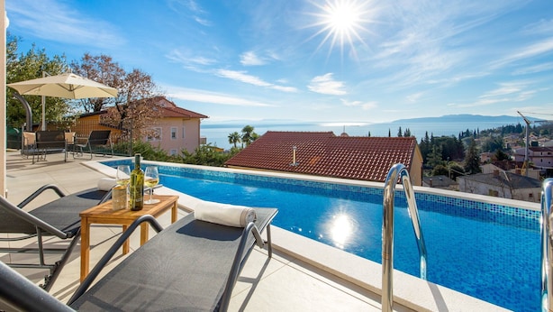 Un espace piscine avec des chaises longues, une table avec des bouteilles et une vue sur la mer et les montagnes.