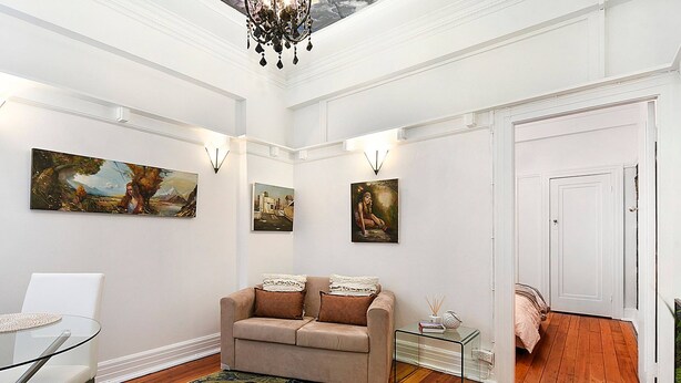 A living room with a beige sofa, a glass coffee table, and a patterned rug. There are three wall-mounted paintings and a chandelier. - ART DECO BAYSIDE APARTMENT CLOSE TO THE CITY (Elizabeth Bay)