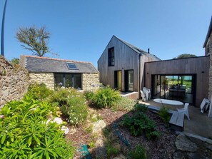 A modern house with a stone wall, a wooden extension, and a garden with plants and a table. - Beautifully renovated house, superb sea view and large garden (Landunvez)