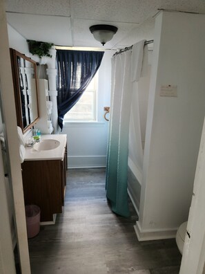 A narrow bathroom with a white sink, mirror, and wooden vanity. A window with a dark curtain allows natural light.