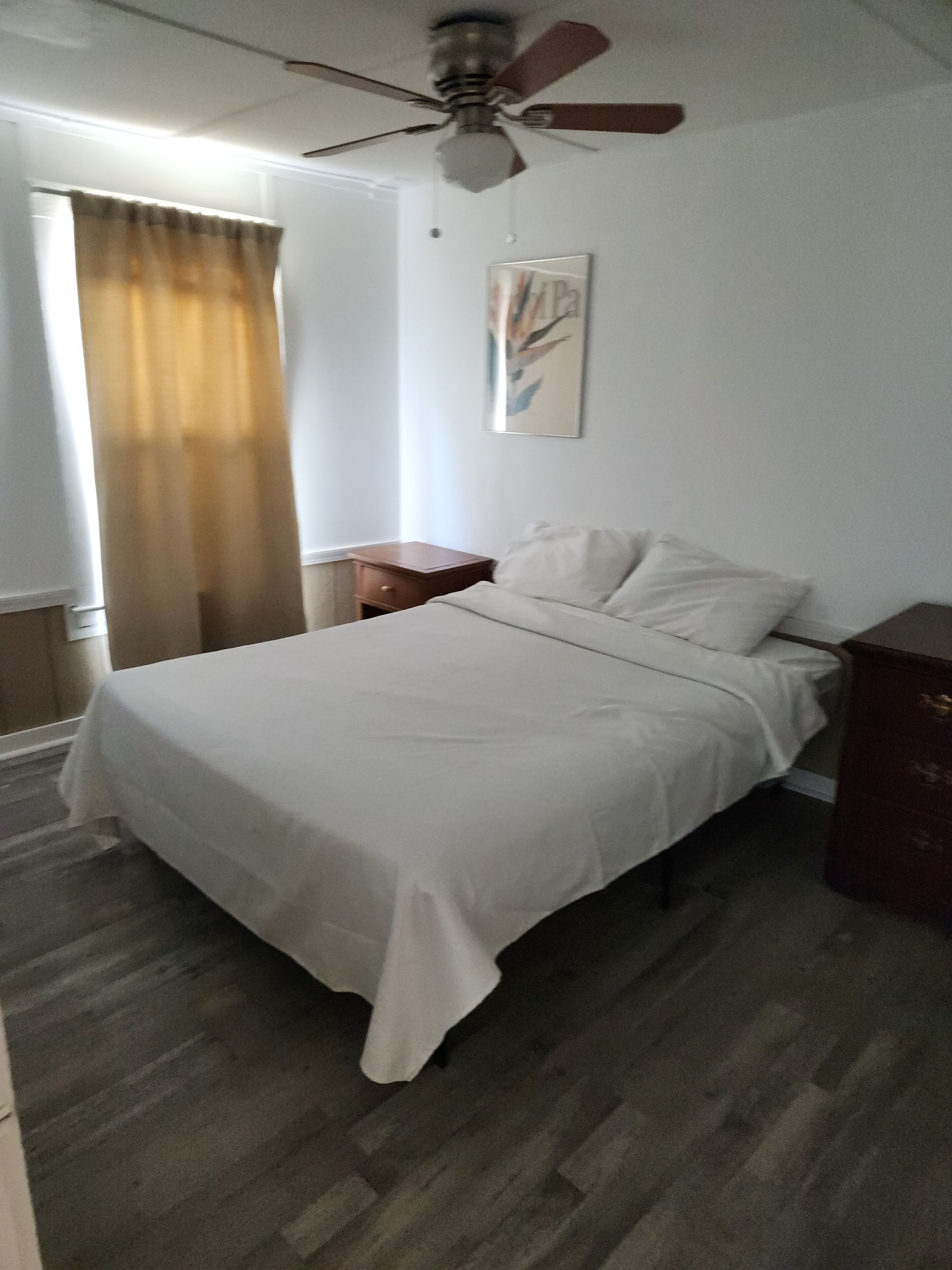 Een slaapkamer met een bed, een plafondventilator, een nachtkastje, een houten dressoir en een raam met gordijnen.