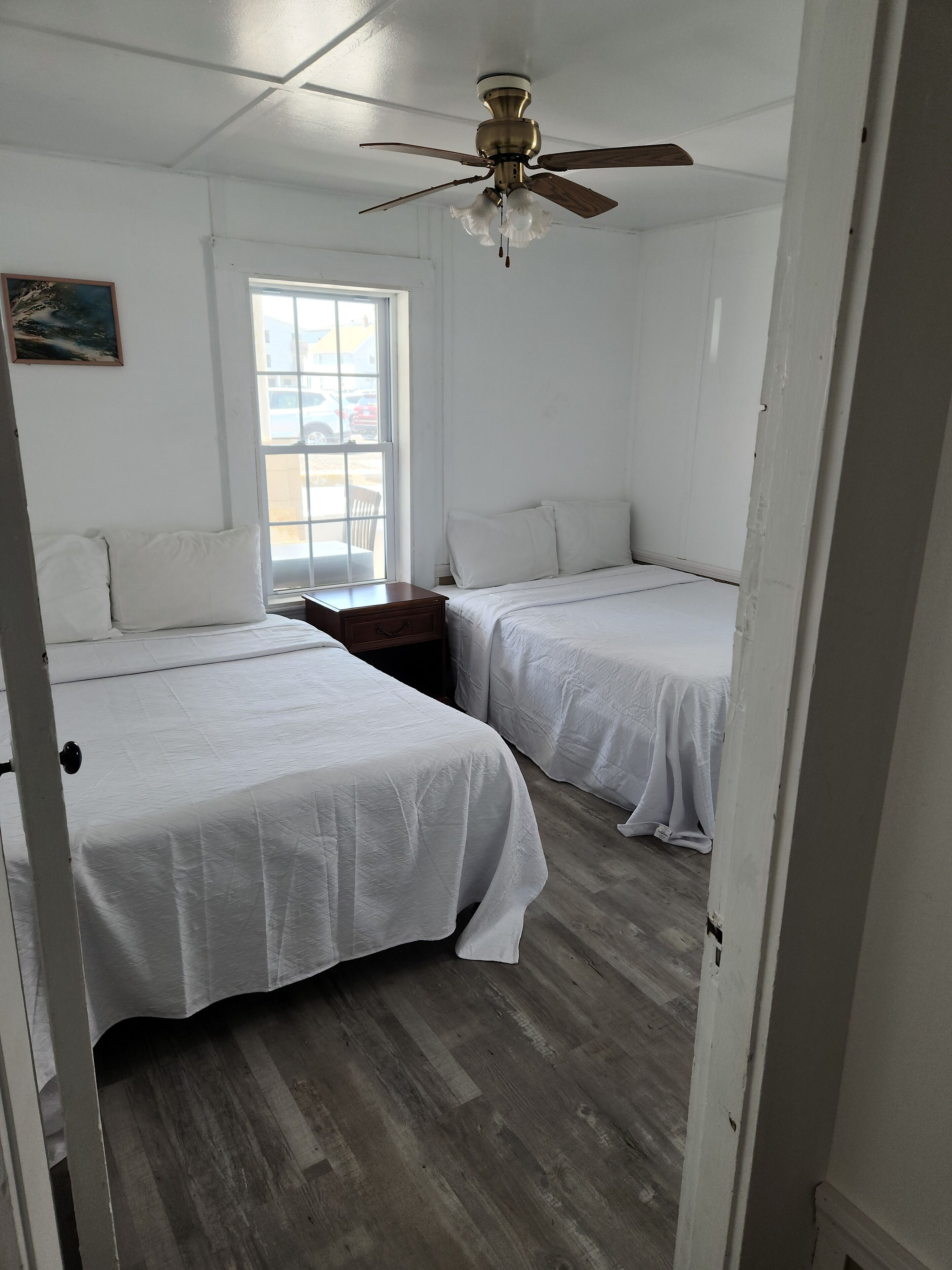 A room with two beds, a ceiling fan, a window, and a painting on the wall.