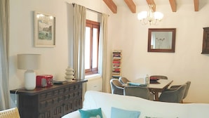 Un salon chaleureux avec un canapé blanc, une table basse en bois et un fauteuil en osier. Trois lampes suspendues sont accrochées au plafond, ainsi qu’une bibliothèque et une fenêtre avec des rideaux.
