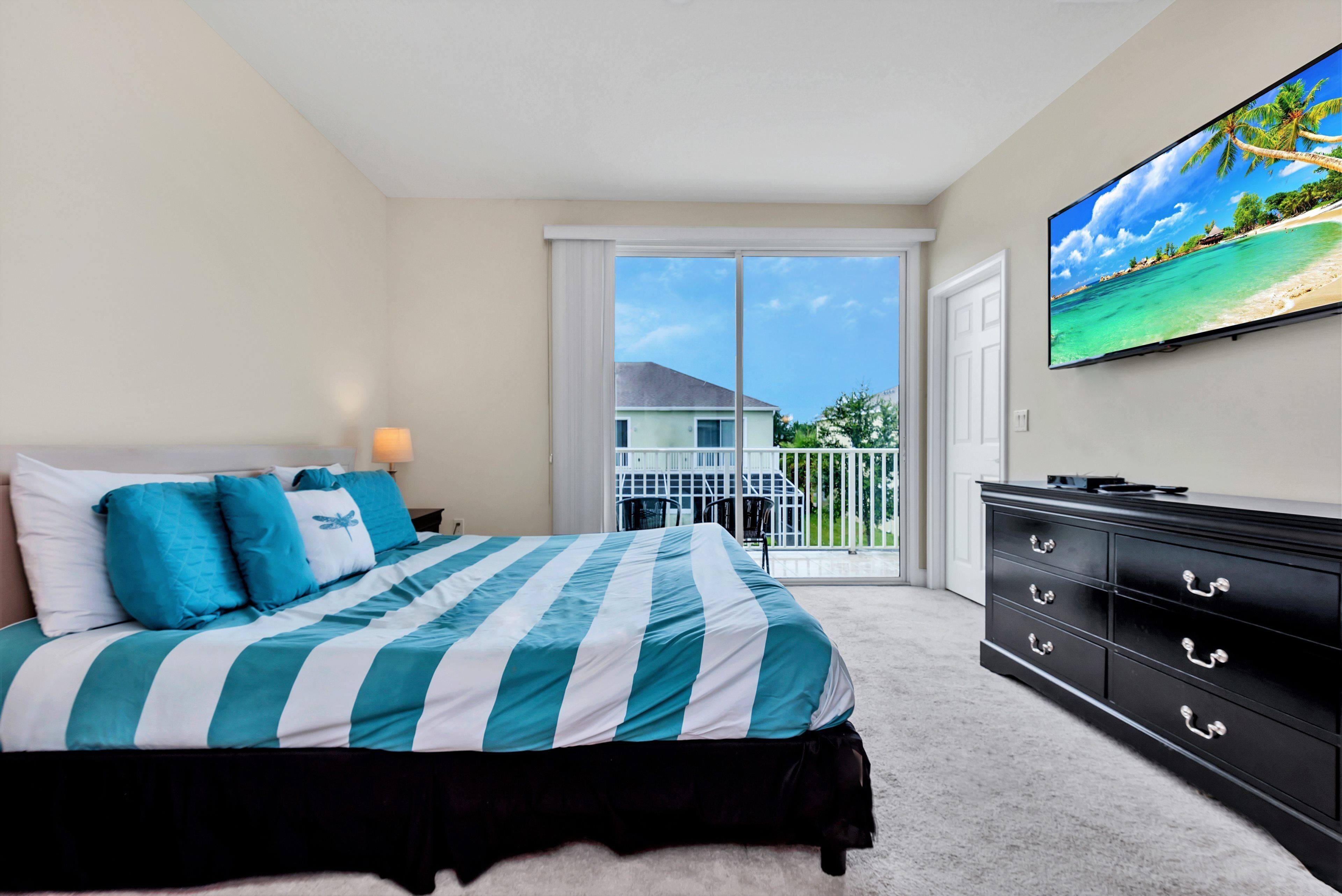 Un dormitorio con cama, televisor, cómoda y balcón con vista a una casa.