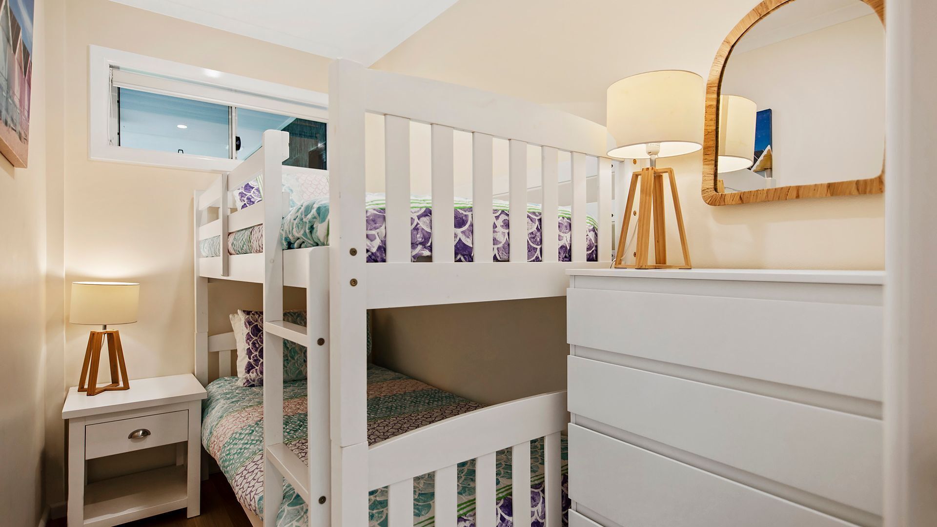 Une chambre d’enfant avec des lits superposés, une table de chevet avec une lampe, une commode et un miroir au mur.