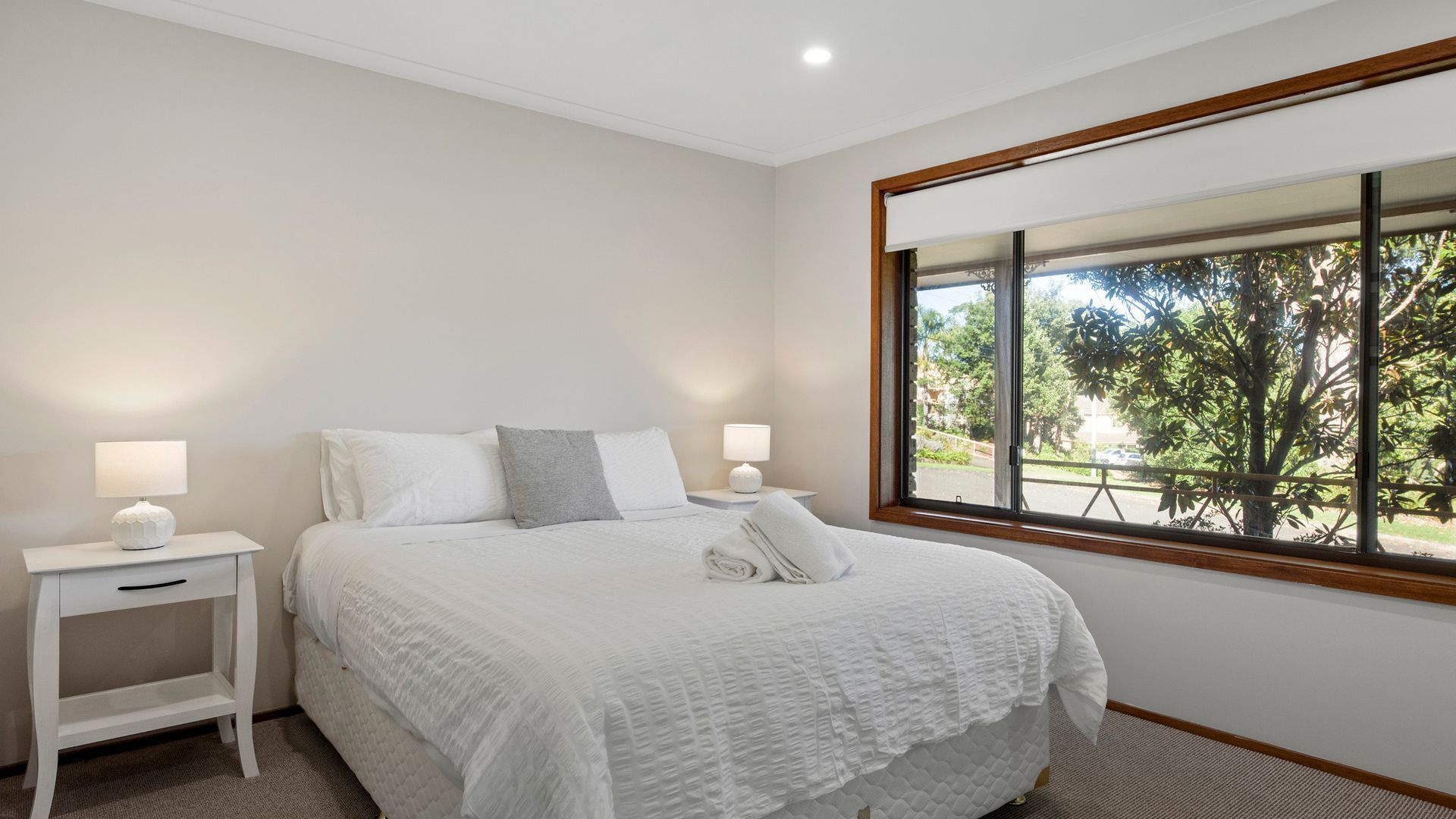 A bedroom with a large bed, two bedside tables with lamps, a window with a view of trees, and a white color scheme.