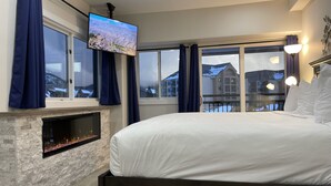 A bedroom with a large bed, a fireplace, a TV mounted on the wall, and a view of buildings outside. - Bright Haus Lakeview at the Village at Breck ,Stunning Views, Ski In/Out In Town (Breckenridge)