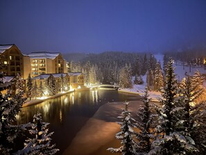Un paysage enneigé avec un lac, des bâtiments dont les fenêtres sont illuminées et des arbres couverts de neige.
